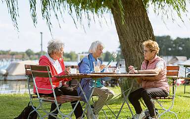 Drei ältere Damen sitzen unter einer Weide am Tisch bei Kaffee und Kuchen. Im Hintergrund ein Fluss mit Booten am Anleger