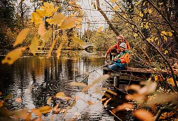 Oma und zwei Enkel sitzen auf einem Steh am Fluss und angeln, umgeben von herbstlichem Wald