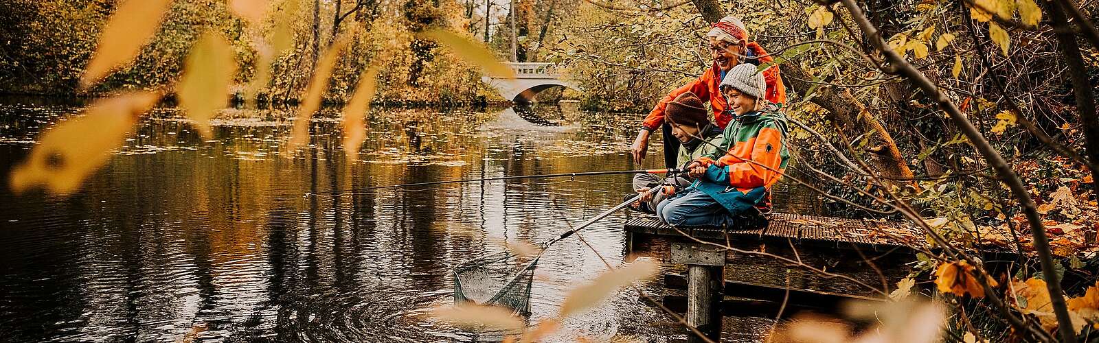 Oma und zwei Enkel sitzen auf einem Steh am Fluss und angeln, umgeben von herbstlichem Wald